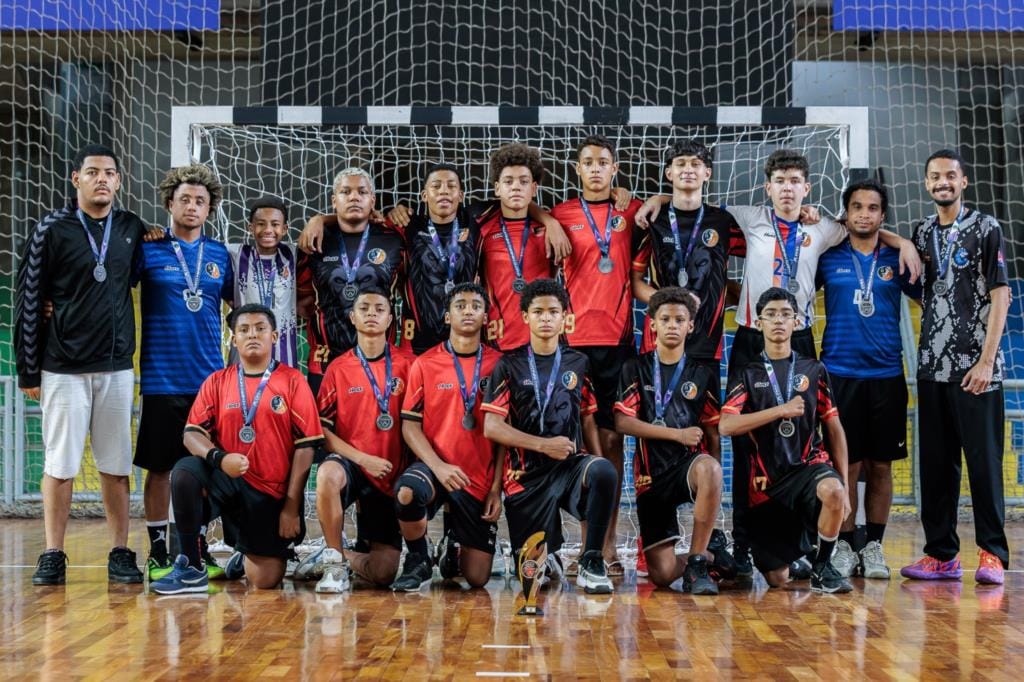 Equipe masculina de handebol de Timóteo posa com medalhas diante de uma baliza, em ginásio esportivo
